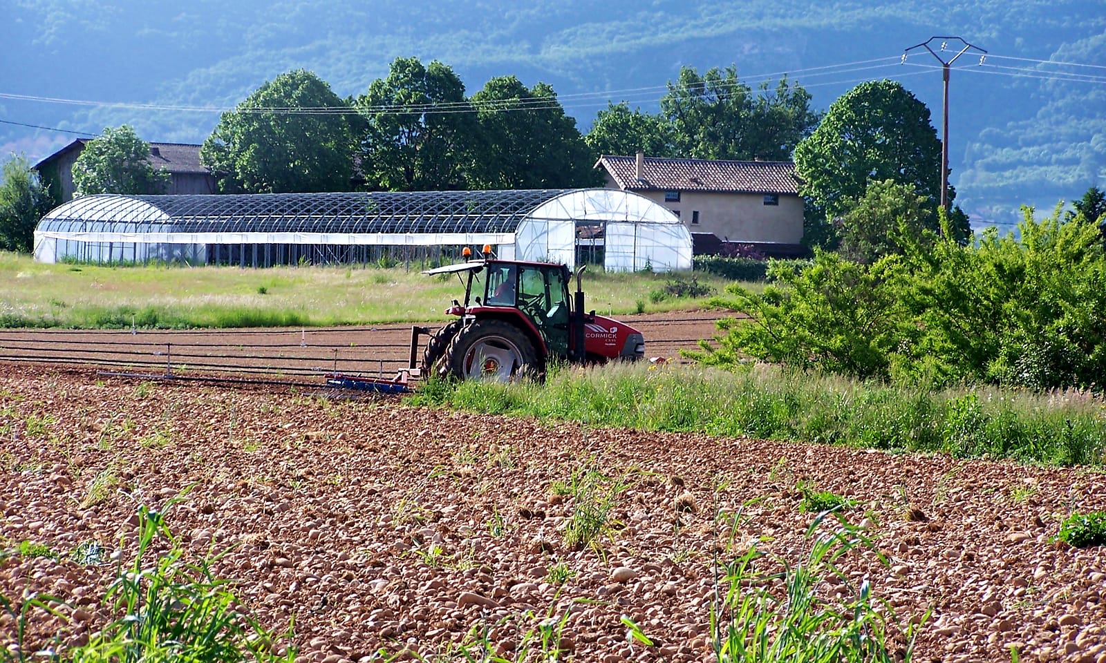 Un tracteur travaille un champ fraîchement retourné, devant les serres d'une ferme et les collines boisées
